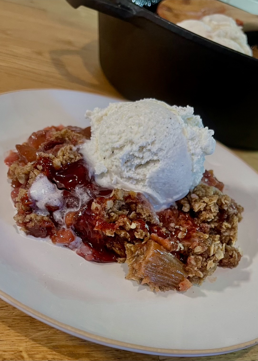 strawberry rhubarb crisp topped with vanilla bean ice cream on a white plate cast iron in the back